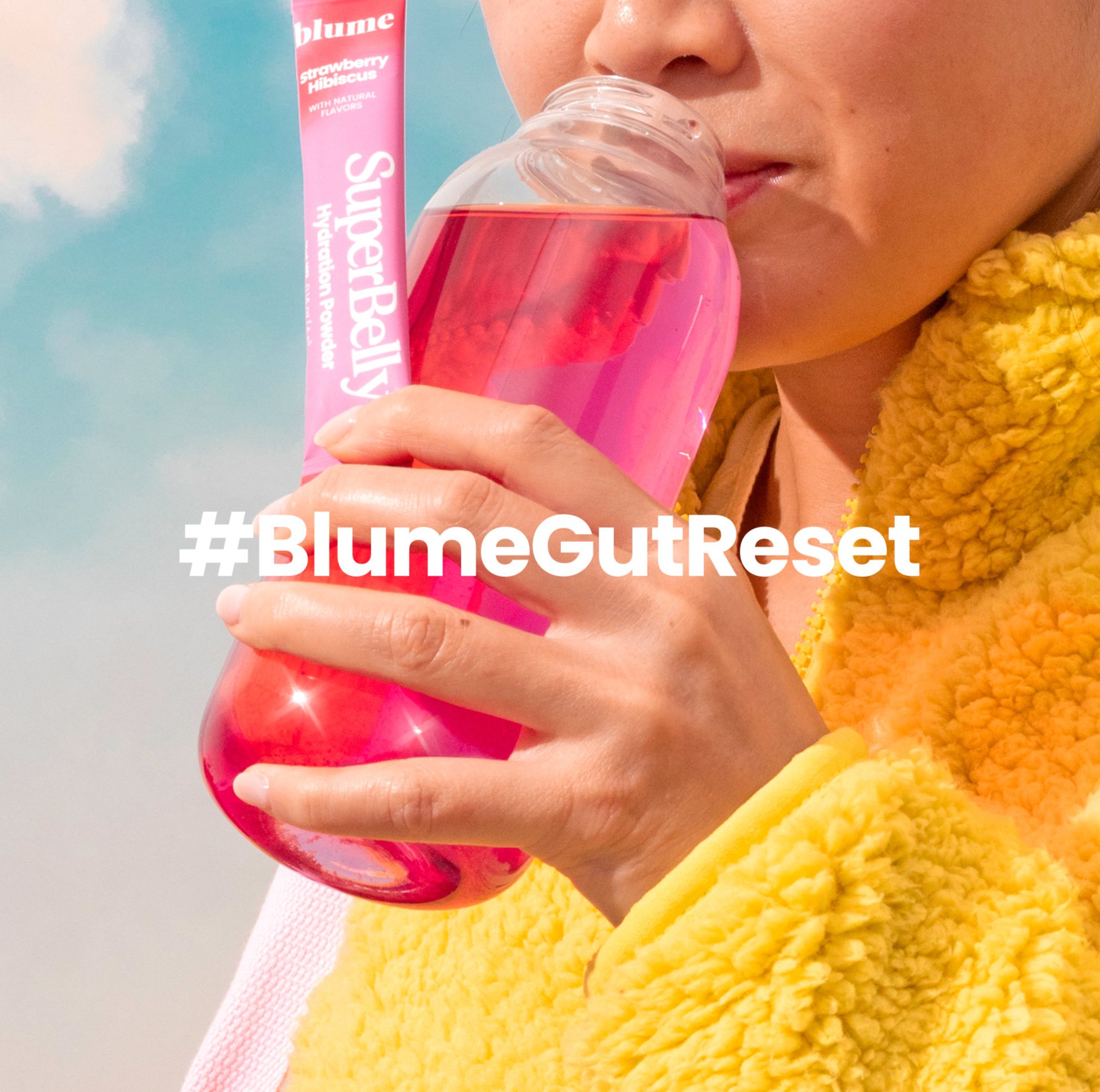 Person in a yellow fleece jacket drinks pink liquid from a bottle, with a pink SuperBelly sachet labeled Strawberry Hibiscus visible. #BlumeGutReset text overlays the image.